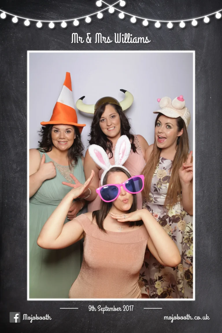 Picture taken by photo booth mirror encapsulated in chalkboard and string lights template. A large group of friends at a wedding reception wearing props and having a good time.