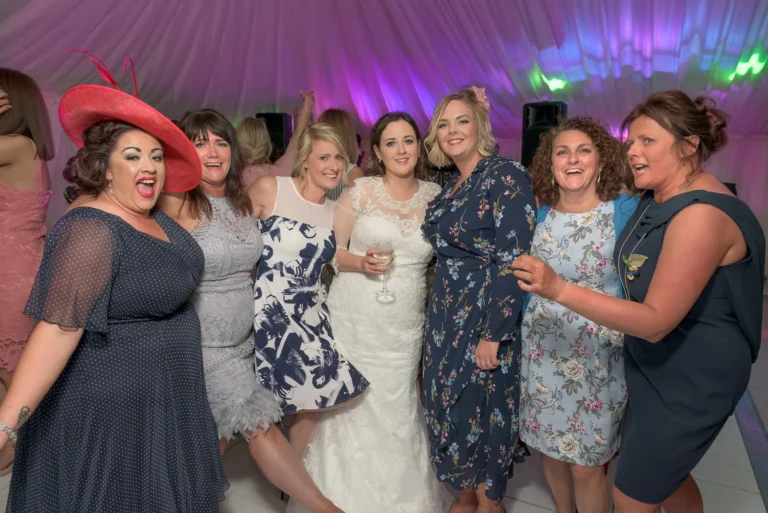 The bride poses with her bridesmaids at a wedding reception. They are in the party mood and having a good time. In the background pink and blue venue uplighting projects on the marquee wall.