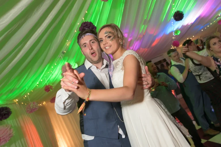 A bride and groom perform the foxtrot at their marquee wedding reception first dance. Multicoloured Venue uplighting accentuates the festival theme they have chosen.